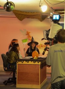 Jade Kalk as a gypsy, Mirriam Meredith as a hippie, and Meghna Bathija as witch sit around the JDTV News anchor desk. Kyle Short is on camera. (Photo by Jeremy Hsieh/ KTOO)