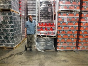 Alaskan Brewing Co. co-founder Geoff Larson stands next to cans of Amber Ale stacked in the Juneau-based brewery’s warehouse. (Photo by Casey Kelly, KTOO - Juneau)