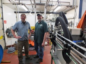 Alaskan Brewing Co. co-founder Geoff Larson (left) and Plant Manager Curtis Holmes stand next to the brewery’s new canning line. (Photo by Casey Kelly, KTOO - Juneau)
