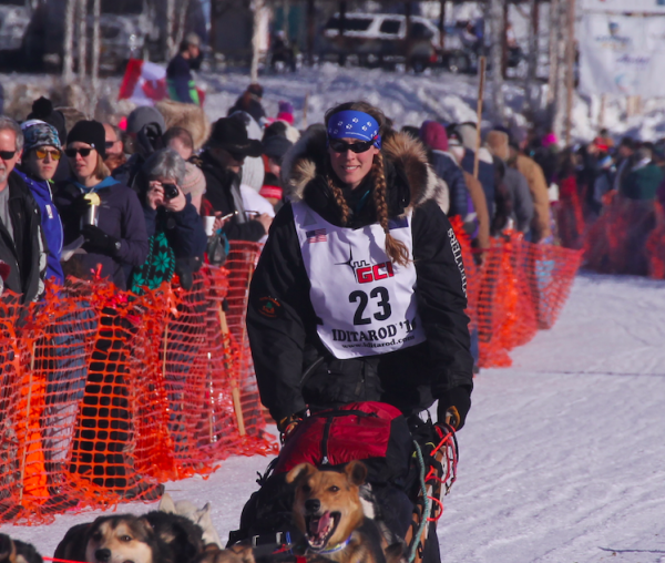 People crowd behind an orange gate to watch mushers and their sled dogs depart on a snow trail.