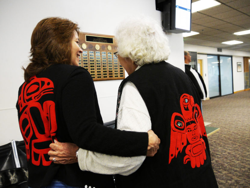 Valerie Hillman shows off her clan’s crest, the T’akdeintaan’s Raven, while holding arms with Hoonah tribal elder Lillian Austin, who’s clan crest is the Shangukeidí’s Thunderbird. The women were on standby to go to Gustavus at Juneau International Airport on Thursday. (Photo by Lakeidra Chavis, KTOO - Juneau)