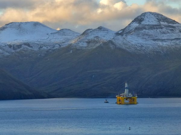 A yellow rig leaves a harbor under snow-covered mountains