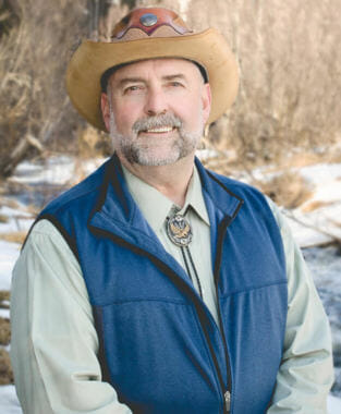A man in a blue vest, a bolo tie, a cowboy hat with a beard