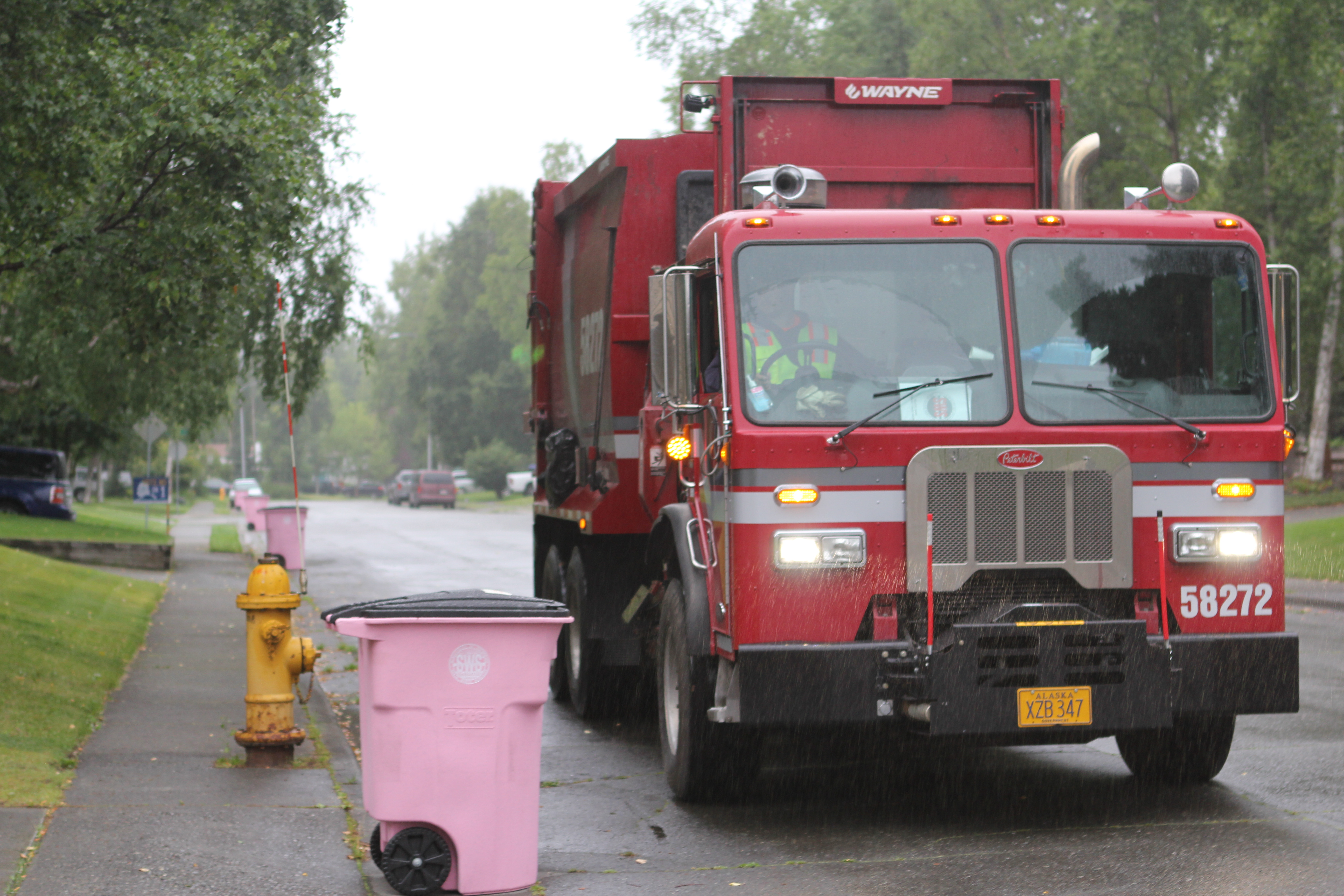 A red trash truck next to a pink trash can on an overcast day