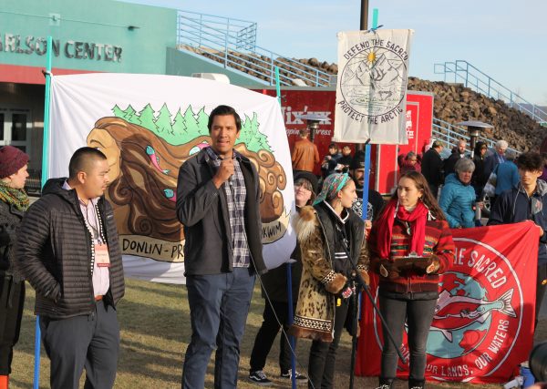 An adult man stands with a group of people speaking into a microphone in front of signs that say "Defend the Sacred"