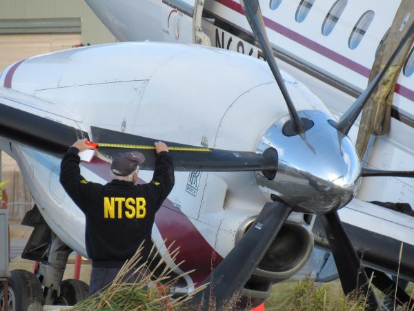 a man in a black uniform with the yellow letters NTSB holds a propellor of a crashed airplane