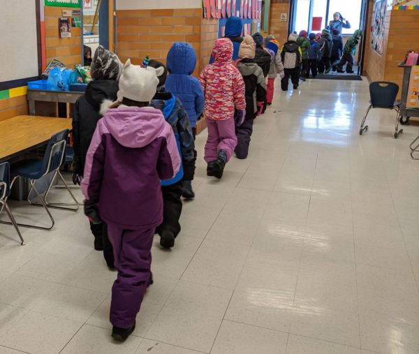 A line of elementary children line up and walk down the hallway out to recess in the winter