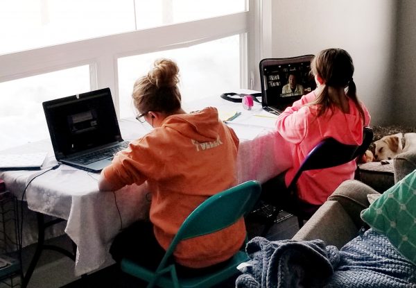 A girl in an orange sweatshirt sits next to a girl in a pink shirt at a desk in their home facing the window while the both do school work on laptop computers