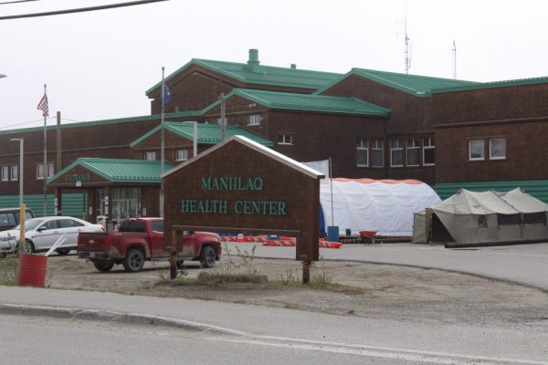 A brown building with a green rood. A red pickup truck is parked in the lot and a sign in green letters says Maniilaq Health Center