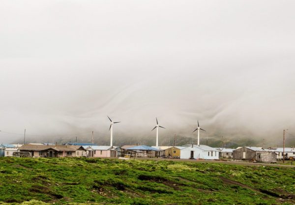 Three relatively small windmills turn above small, one-story homes. Green grass in the foreground and thick fog in the background .