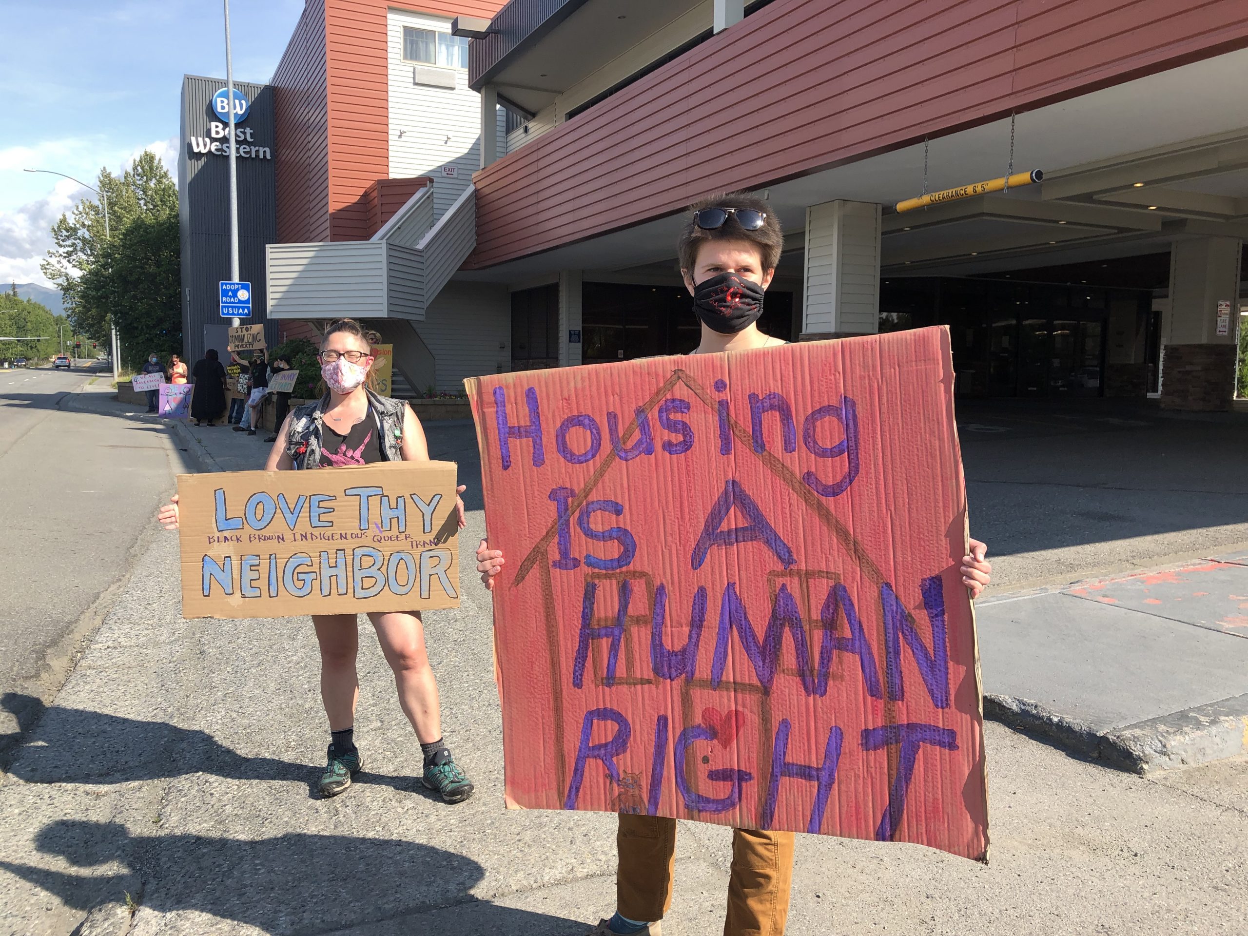 Two people wearing masks hold protest signs on a sidewalk in front of the Best Western Golden Lion. One sign says "Love thy neighbor," and the other says "Housing is a human right."