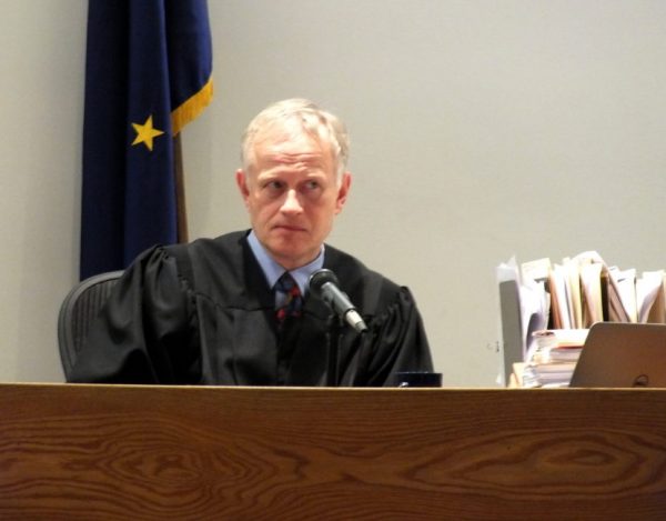 A white old man in a judge's robee sits at a desk in front of an Alaskan flag