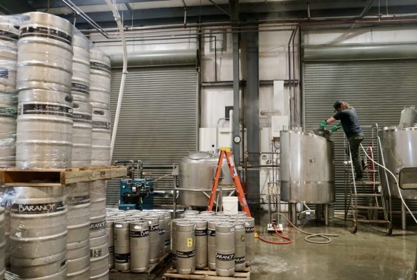 Stacks of silver canisters and kegss sit around a crowded warehouse along with ladders, tools, and a garage door in the back. 