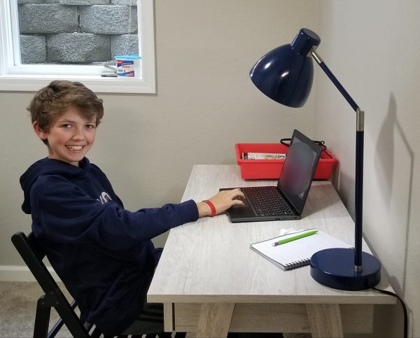 A young boy sits at a desk and smiles at the camera while working on a laptop