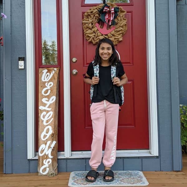 A young girl stands in front of a front door to a house smiling and wearing a back pack