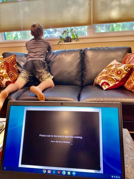 A young boy kneels on a couch and looks out the window as a laptop screen behind him shows a loading page