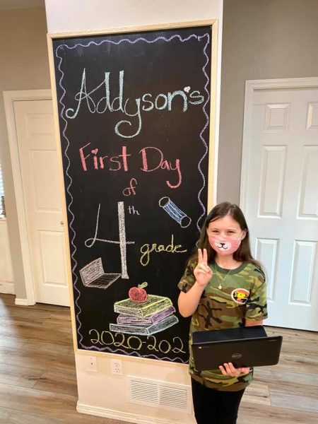 A young girl puts up the peace sign with one hand and holds a laptop in the other in front of a sign that says 'Addyson's first day of 4th grade'