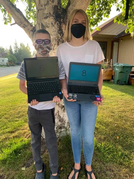 A boy and a girl stand in front a tree wearing face masks and holding laptops