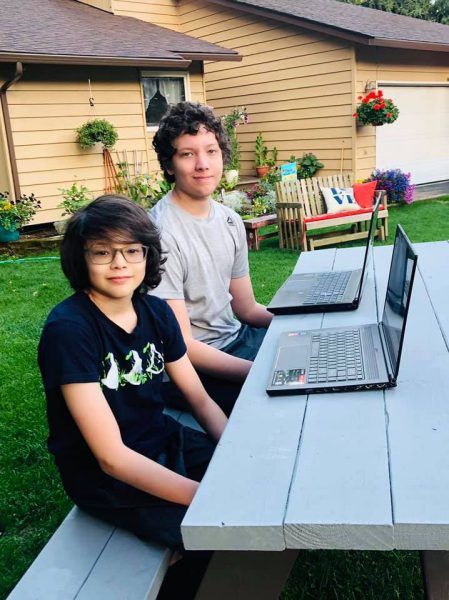 Two boys sit at a picnic table outside with laptops