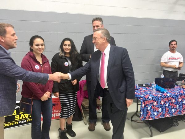 A man reaches across the frame to shake hands with another man in a school cafeteria. Political placards are in the background, as is one man wearing a "Mike Dunleavy for governor" t-shirt.
