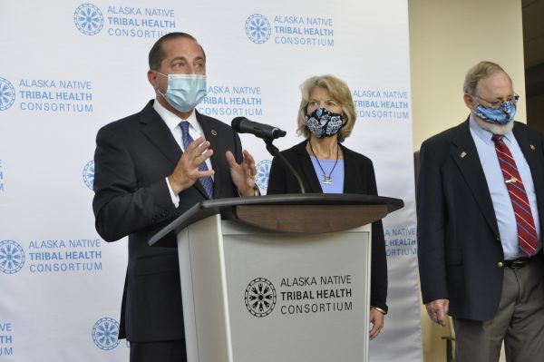 A man in a suit stands at a podium that says on it "alaska Native Tribal Health Consortium," as does the backdrop. To the viewers right is Sen. Lisa Murkowski and Rep. Don Young. All three are wearing masks.