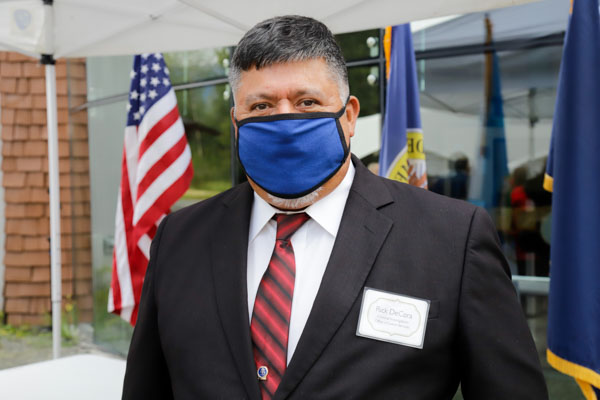 A big man in a suit an tie with a blue face mask stands in front of a U.S. flag with other flags in the background.