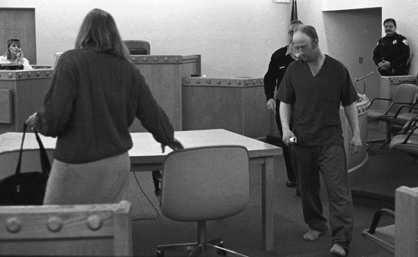 In a black and white photo, Richard Bingham walks towards a chair as a woman whose back is turned pulls ist out. 