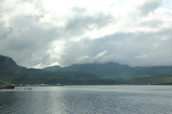 Clouds hang over forested bay, where some boats and buildings are visible in the distance. 