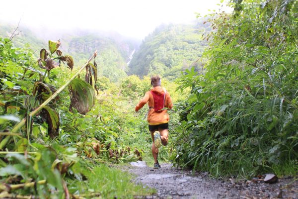 A runner bounds down a wet dirt trail wearing black shorts, a red backpack and an orange windbreaker. Mountains rise into mist in the background and there are wet leaves and other vegetation in the foreground.