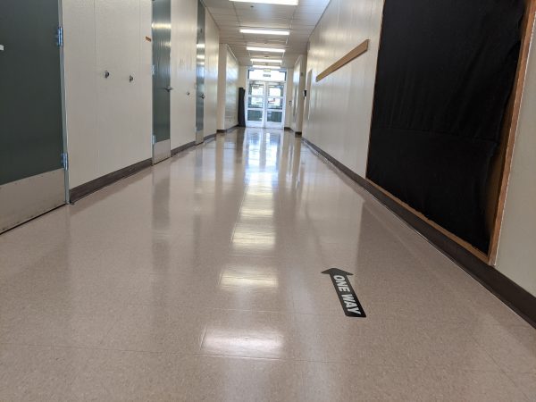 An empty hallway in an elementary school with a black arrow sticker on the floor pointing in one direction that says "one way"