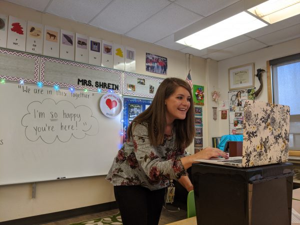 A woman smiles and bends over to look at her laptop screen in her 5th grade classroom