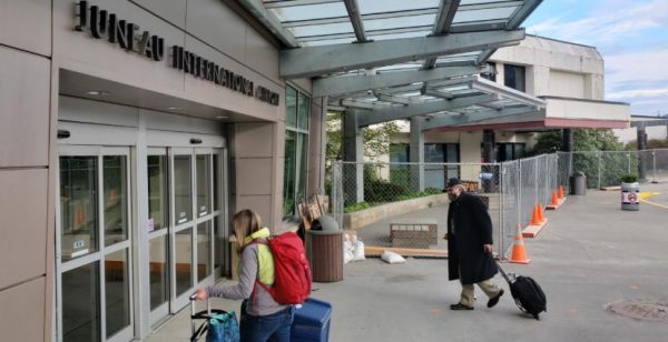 Two people carry luggage towards the sliding glass doors under an awning at the Juneau Airport