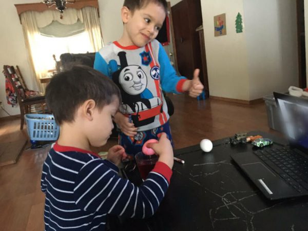 Two boys play with toys on a table at a home. One boy is weraing a blue train shirt and is giving a thumbs up.
