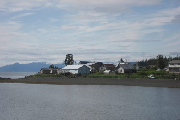 A photo taken from the water of a handful of painted wooden houses and a church steeple with mountains in the backgrorund.