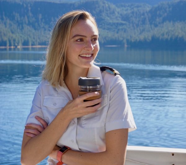 A woman in a white shirt with black epaulettes holds a cup and smiles. She's on a boat deck in calm water. Forested slopes are in the background.