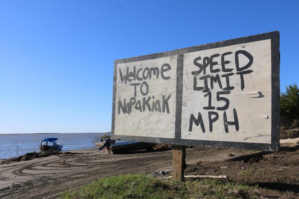 A hand-painted piece of plywood stands over lookign the water. It reads "Welcome to Napakiak - Speed Llimit 15 MPH"