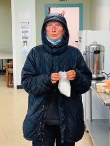A woman in a black raincoat with a blue surgical mask over her ears stands next to a coffee carafe