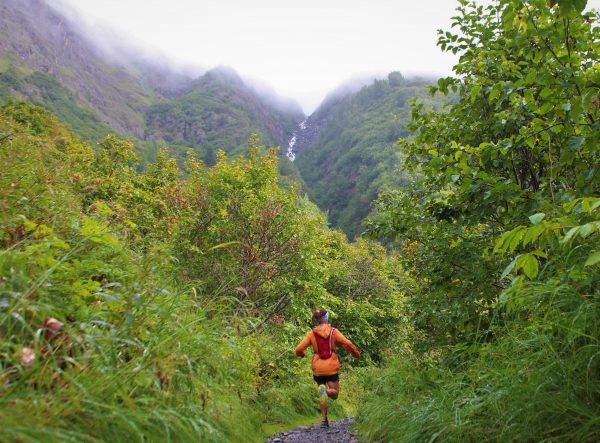 A runner in an orange windbreaker and wearing a red running backpack descends a dirt trail with wet vegetation in the foreground and mountains rising into fog int he background