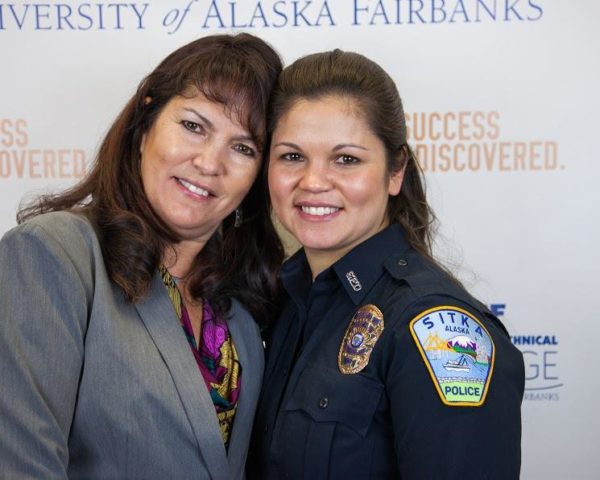 Two brown haired women, one in a gray suit and one in a blue police uniform