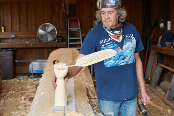 A man in a blue tshirt, bandana, with gray hair and a goattee holds a sculpture of a hand holding a feather in a wood shop