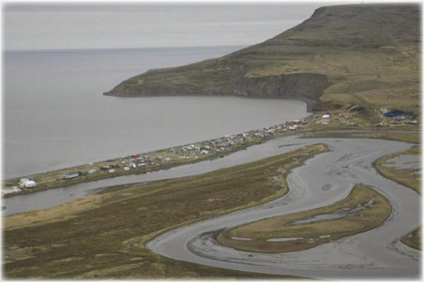 An aerial image shows a winding river delta next to a bluff. Several dozen wooden houses stand on the berm abutting the water. 