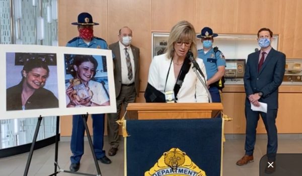 A white woman with blond hair in a white suit and an arm in a black sling speaks on a podium with the DPS logo and two photos of a teenage girl on an easel on the lefthand side of the image