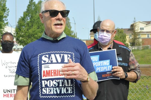 A man in a blue t-shirt and sunglasses. His t-shirt reads "Save America's Postal Service." Behind him are people wearing masks holding placards.