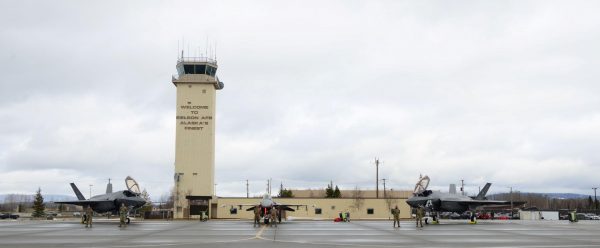 Two f-35 fighter jets are parked in front of a beige aircraft control tower 
