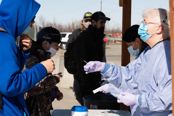 A man in a blue hospital gown and a blue surgical mask passes a swab to a man in a blue hoody from across the table