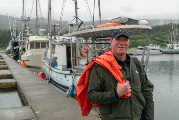 Al Gross stands on a boat dock holding fisherman's jacket over his shoulder, looking at the camera