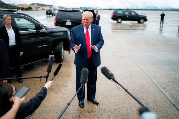 Donald Trump, wearing a black suit and dred tie, gestures in a rainy lot next to several black SUVs with microphones visible in the foreground