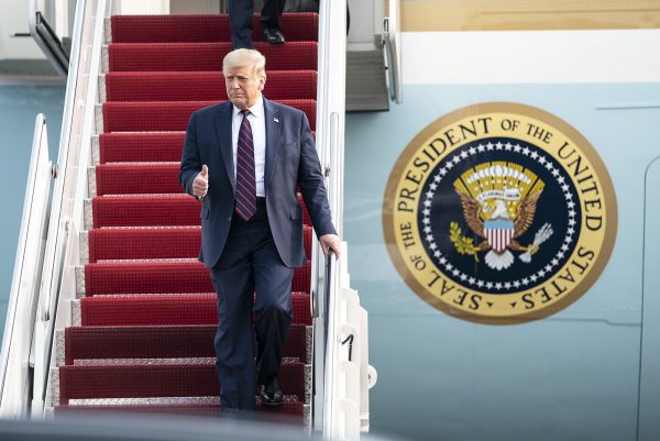 President Trump, wearing a black suit andd giving the thumbs up, descends from the red stairs of air force one with the presidential seal next to him. 