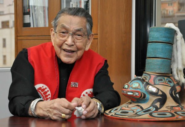An elder Alaska Native man in a red vest smiles while sitting at a table