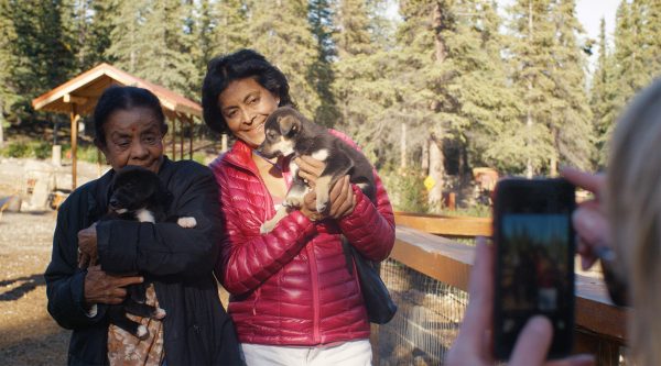 Two women hold puppies and pose for a smartphone photo in front of spruce trees